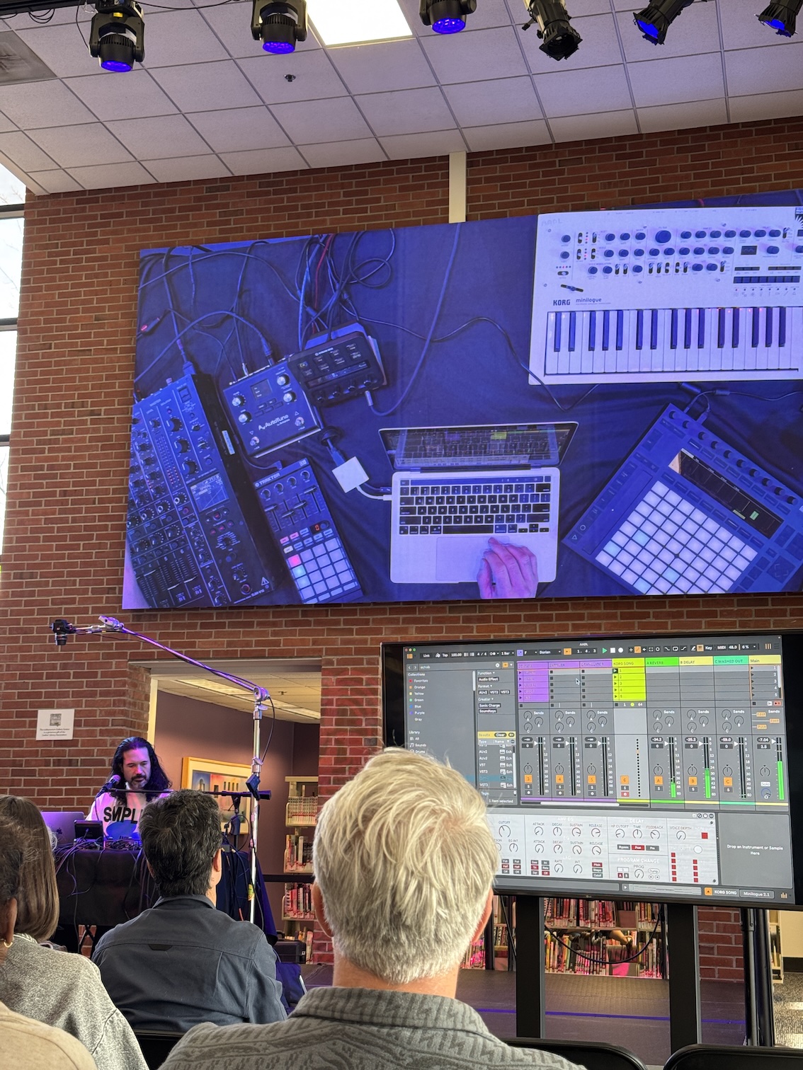 A man standing on stage standing in front of a projector screen shows the audience how he makes music on Ableton software.