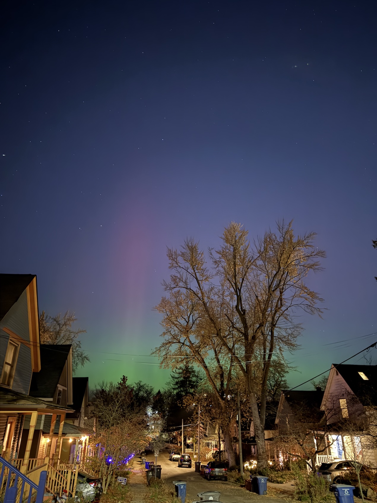 A beautiful blue, green, and purple aurora streaking through the night sky above a dimly-lit neighborhood street